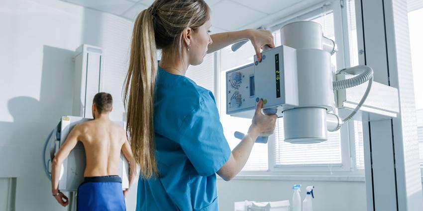 The image shows a healthcare professional adjusting an X-ray machine while a patient stands ready for the scan, highlighting a health monitoring or diagnostic procedure in a medical setting.
