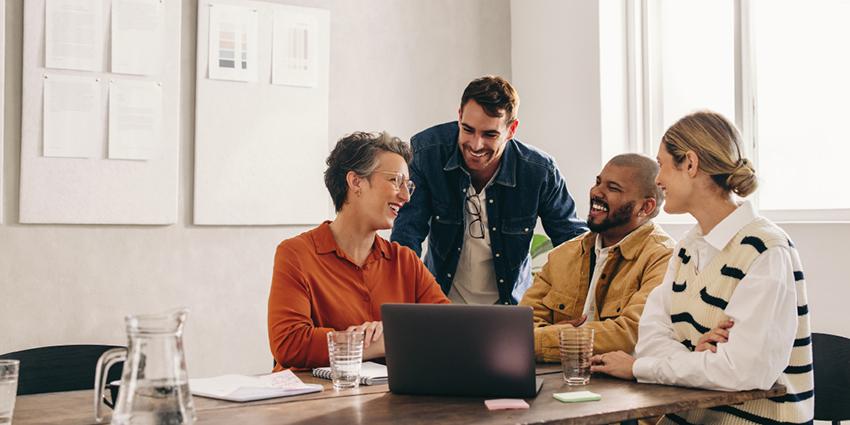The image shows a group of colleagues gathered around a table with a laptop, smiling and engaged in discussion