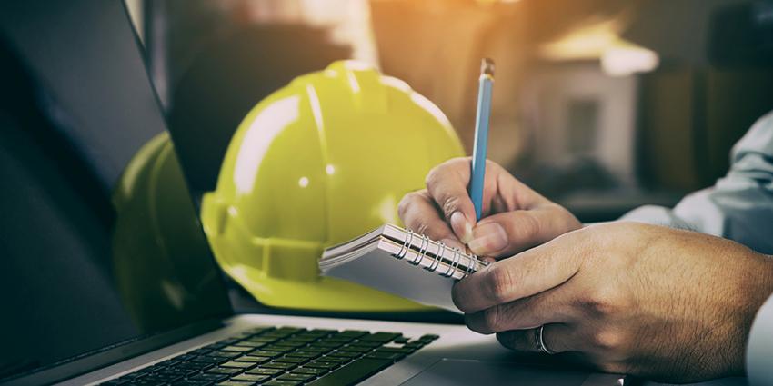 The image shows a person taking notes on a notepad beside a laptop, with a yellow hard hat in the background, suggesting a focus on workplace safety or reporting tasks.
