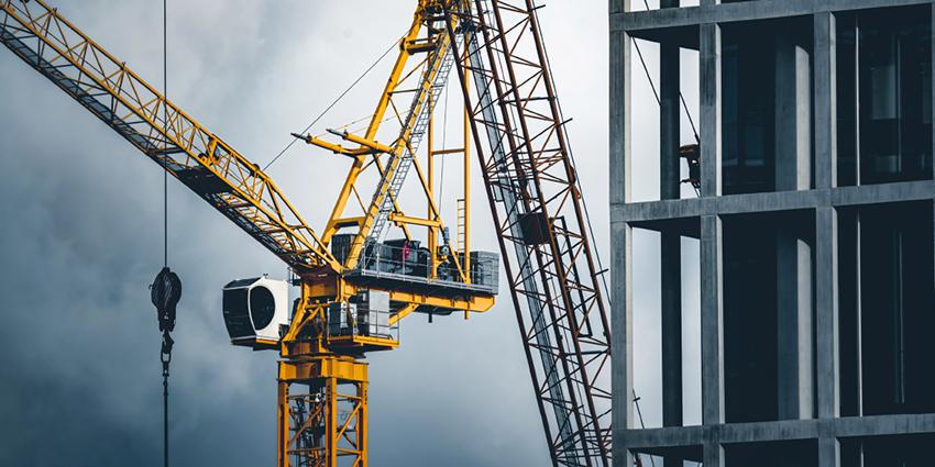 The image shows a construction site with large cranes next to a multi-story concrete building under a cloudy sky, emphasizing heavy equipment and structural development.