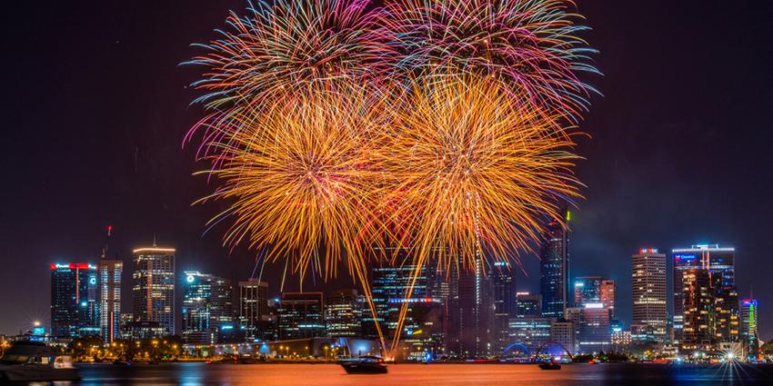 The image shows vibrant fireworks lighting up the night sky above a city skyline, with reflections on the water